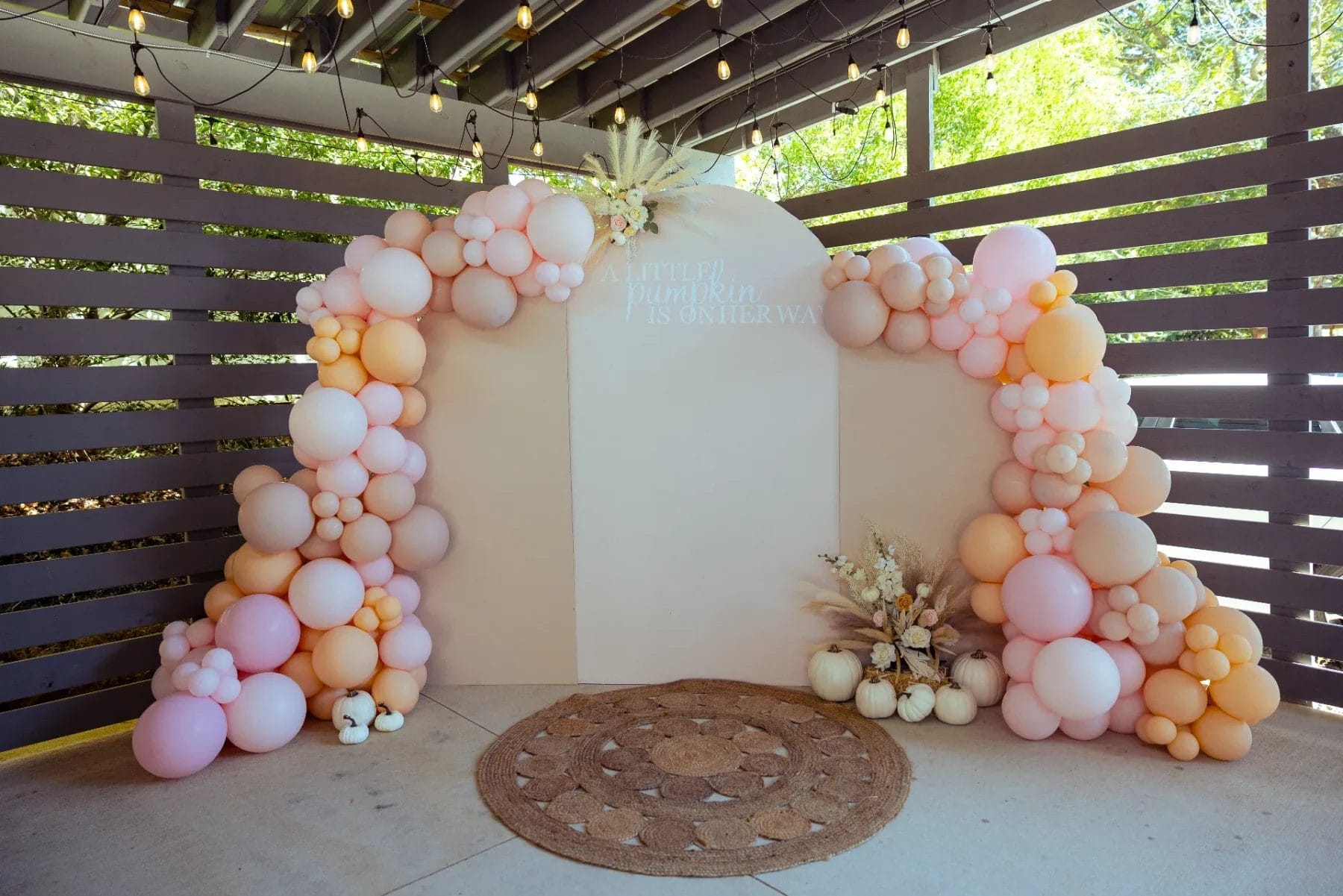 A decorated photo backdrop under a pergola, featuring clusters of pink, peach, and cream balloons. Dried flowers and pumpkins sit at the base, with a rug in front. The sign reads "A beautiful pumpkin is on her way. Huntersville event venue