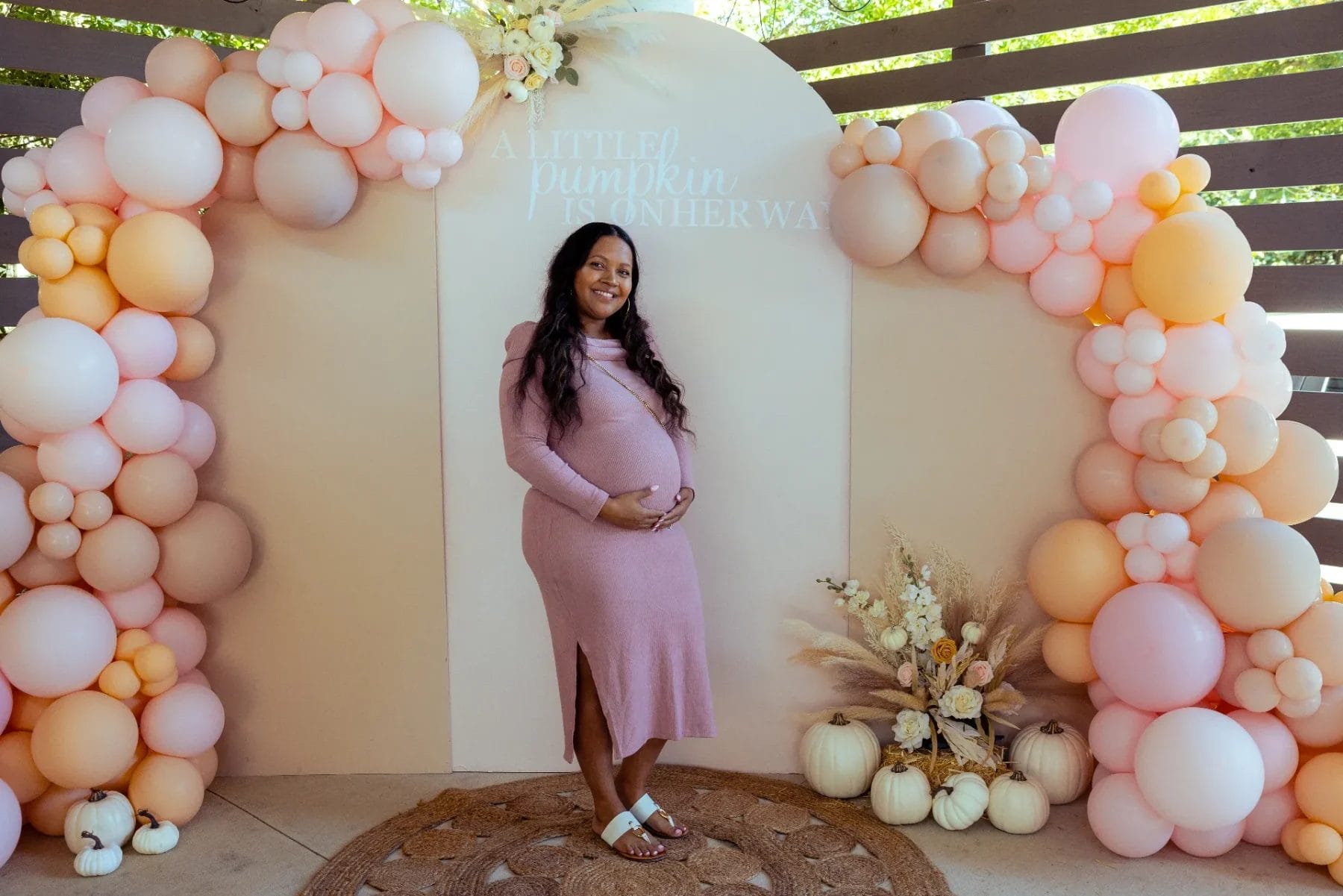 A pregnant woman in a pink dress stands smiling in front of a decorative arch made of pink and peach balloons. There's a sign above her that reads "A Little Pumpkin is on Her Way." Pumpkins and flowers decorate the area. Huntersville event venue