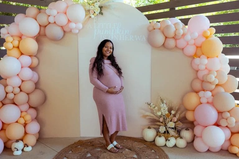A pregnant woman in a pink dress stands smiling in front of a decorative arch made of pink and peach balloons. There's a sign above her that reads "A Little Pumpkin is on Her Way." Pumpkins and flowers decorate the area. Huntersville event venue