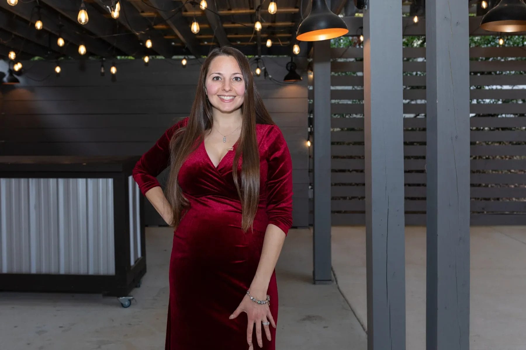 A woman in a red velvet dress stands smiling under string lights on a patio with dark wooden walls and metal support beams. She has long brown hair and her hand rests on her hip. Huntersville event venue