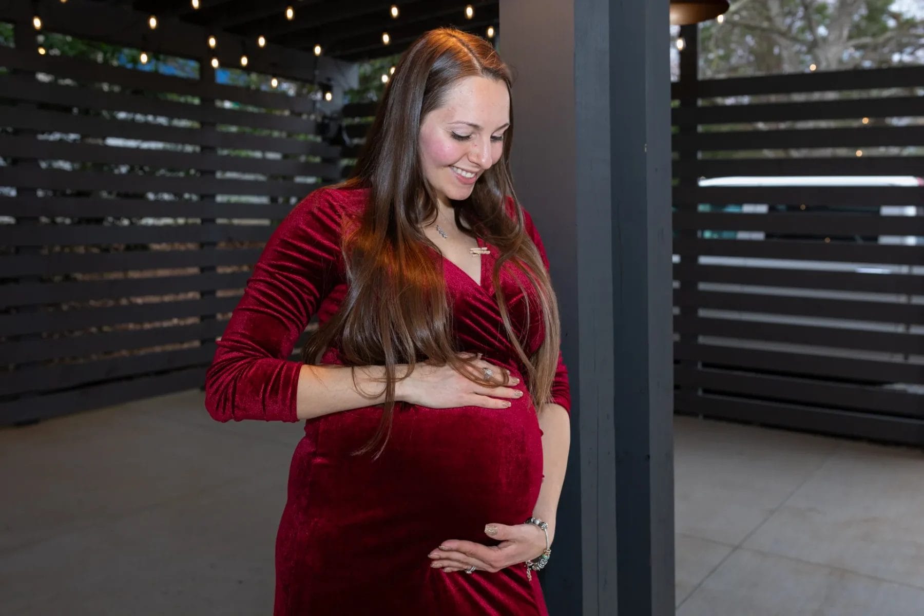 A smiling woman in a red velvet dress stands beneath string lights, gently holding her pregnant belly with both hands. She is surrounded by a modern outdoor setting with wooden paneling. Huntersville event venue