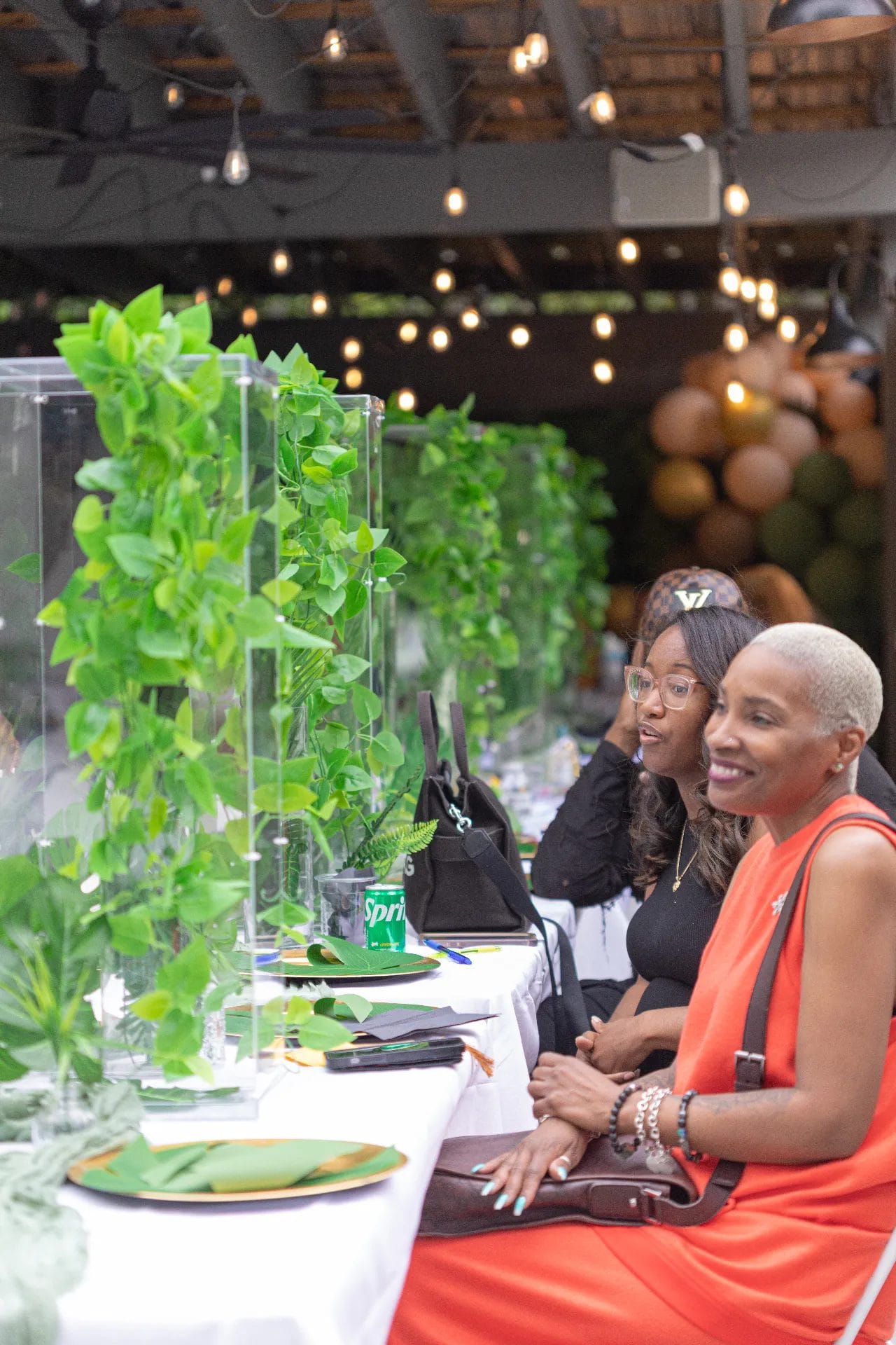 Three women sit at a decorated table with green foliage centerpieces at an outdoor event. The woman in front wears an orange outfit and smiles. Others are engaged in conversation. Balloons and string lights decorate the background. Huntersville event venue