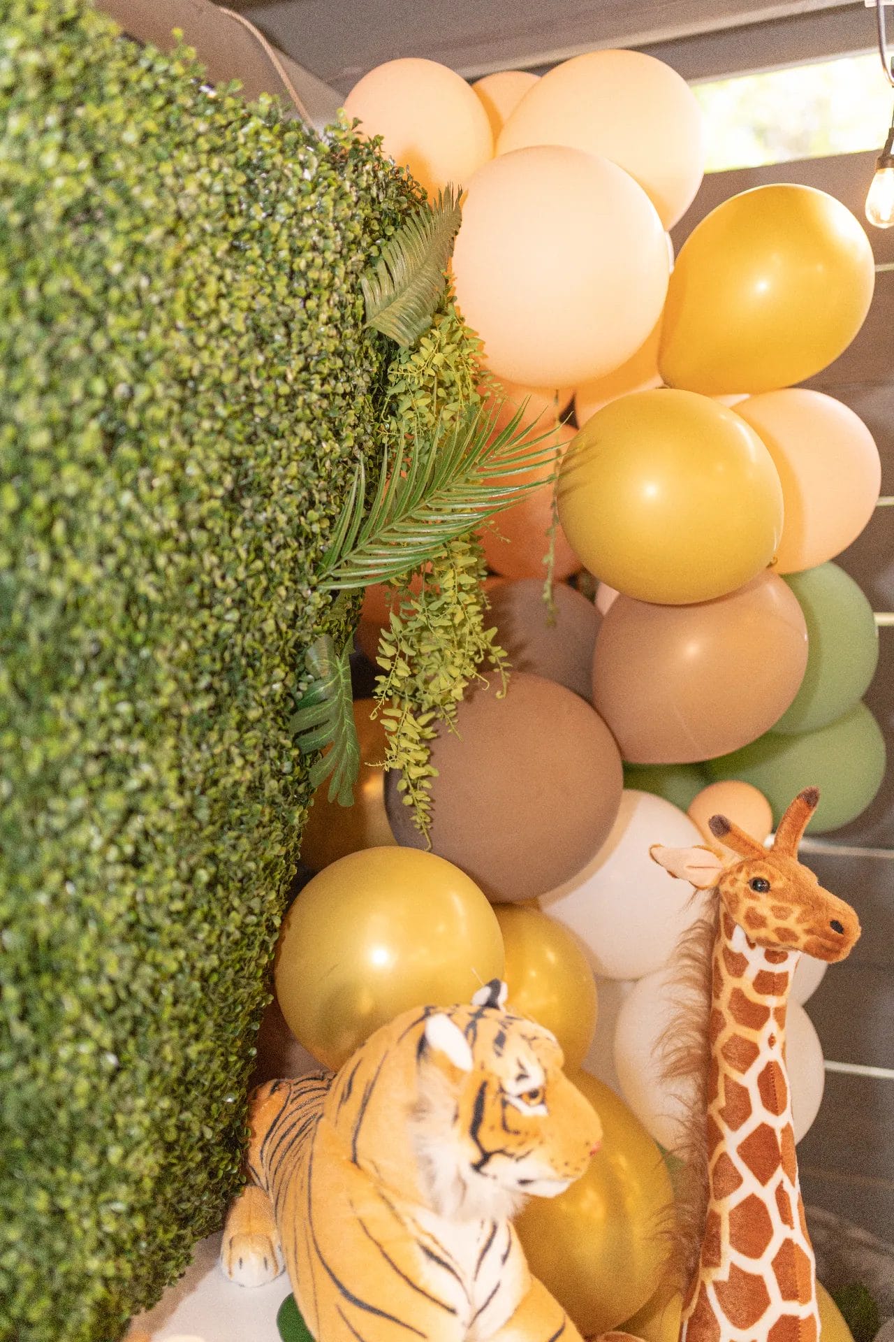 A decorative setup features a lush green hedge, clusters of yellow and brown balloons, and plush toy animals, including a giraffe and a tiger, adding a lively touch to the scene. Huntersville event venue