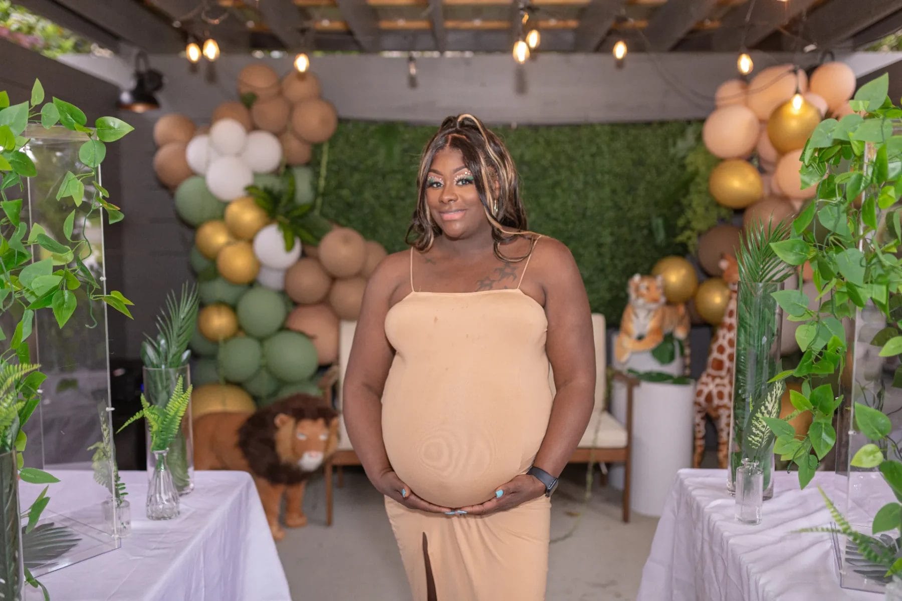 A pregnant woman stands smiling at a baby shower. She is wearing a beige dress, and the background is decorated with green, brown, and gold balloons, greenery, and plush animals, creating a jungle theme. Huntersville event venue