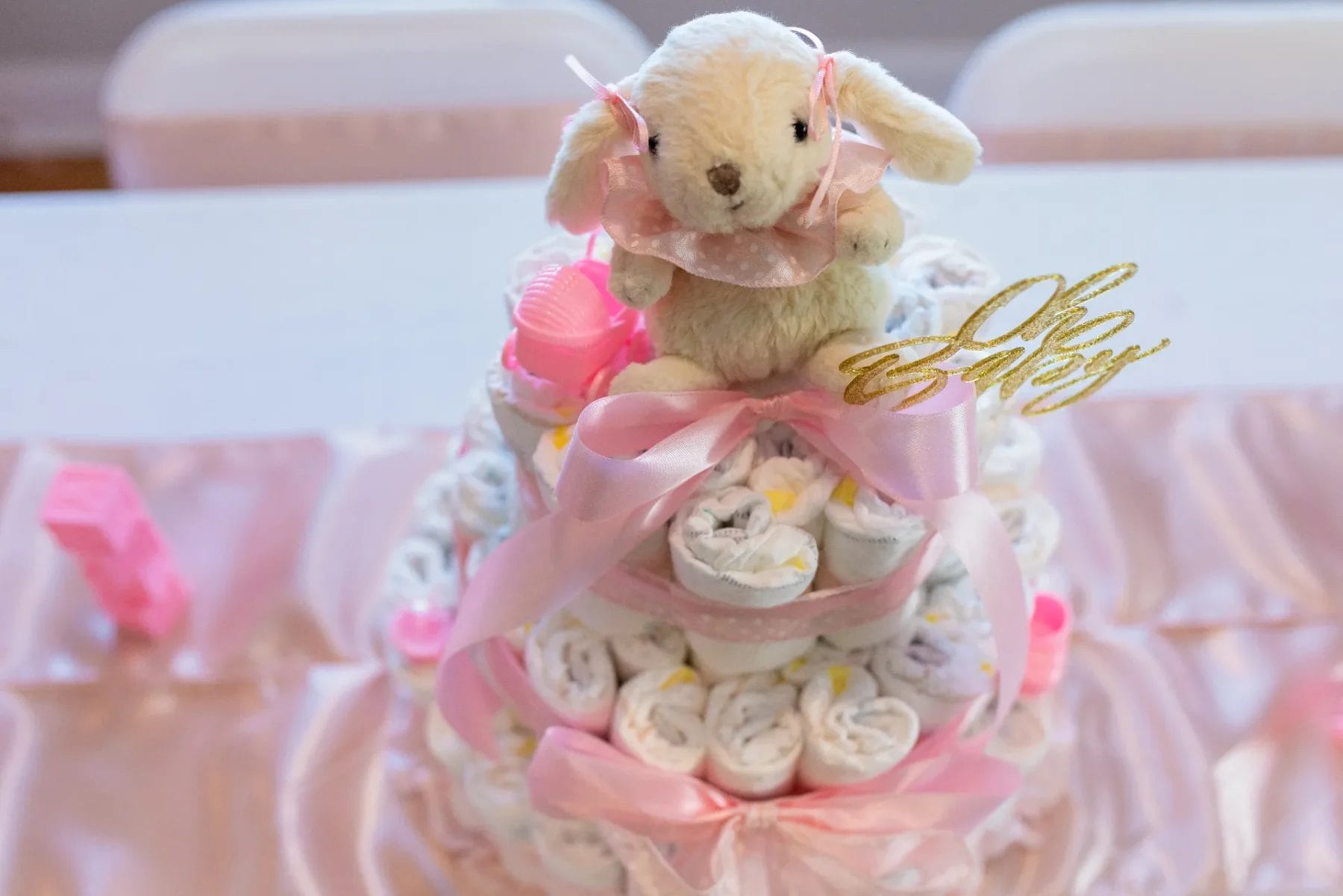 A diaper cake decorated with pink ribbons and topped with a stuffed lamb. Gold "Oh Baby" sign attached. Placed on a pink satin tablecloth with white chairs in the background. Huntersville event venue