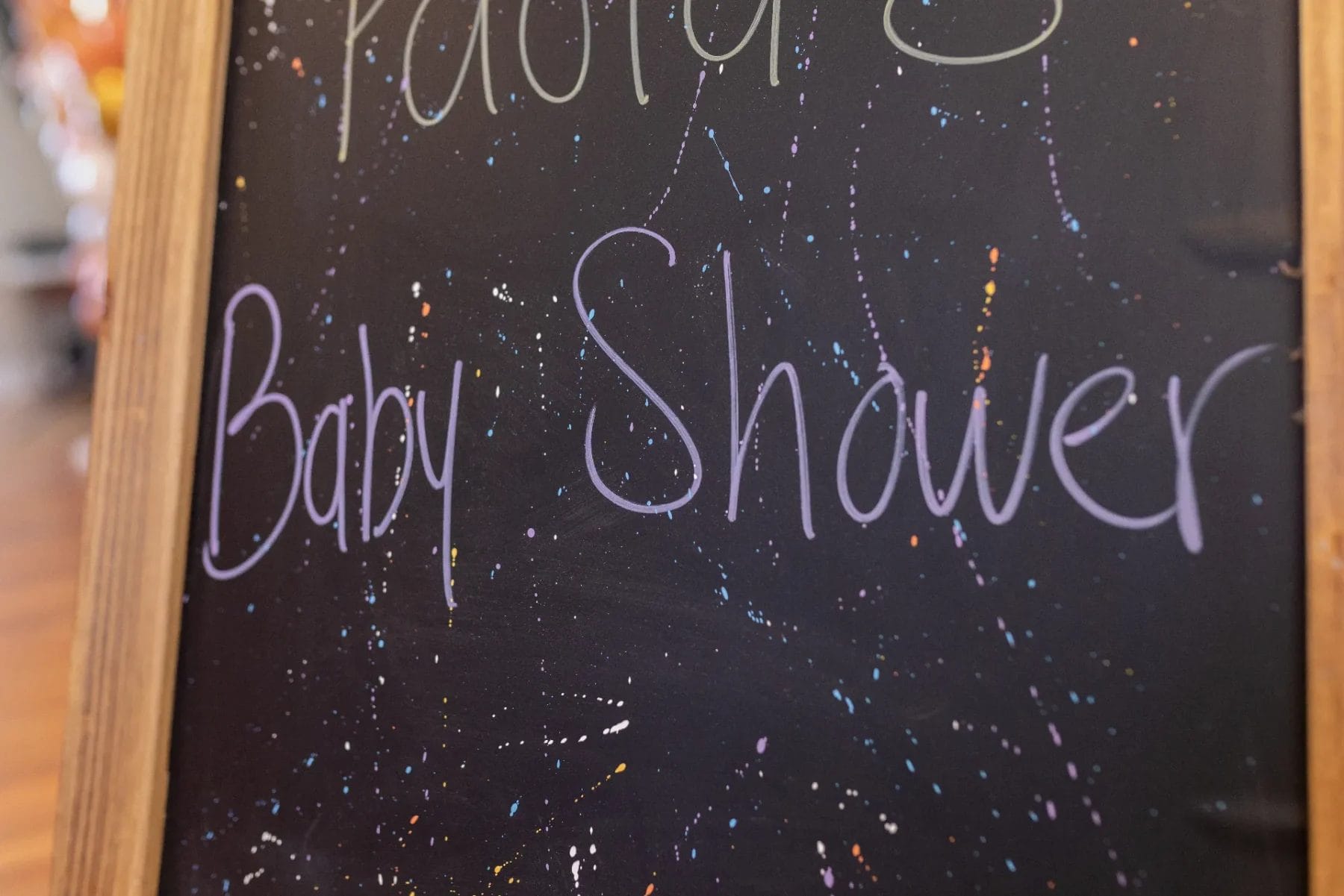 A chalkboard sign with colorful splatter design reads "Baby Shower" in purple handwriting. The board has a wooden frame and is standing indoors on a wooden floor. Huntersville event venue