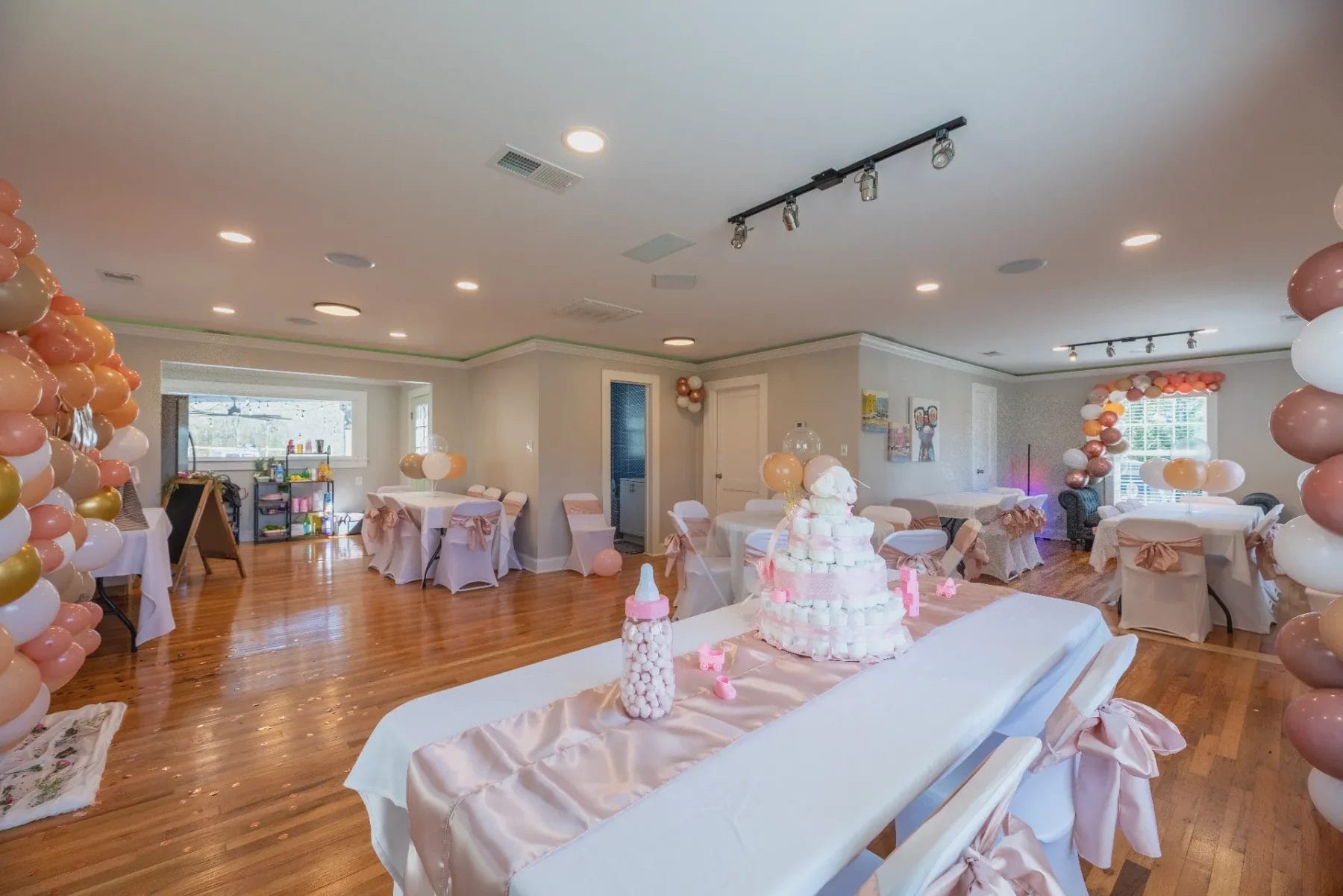 A well-decorated room for a baby shower features pink and white balloons, tables with white cloths and pink bows, and a diaper cake centerpiece. A large baby bottle decoration is on one table, with gifts stacked on another table in the background. Huntersville event venue