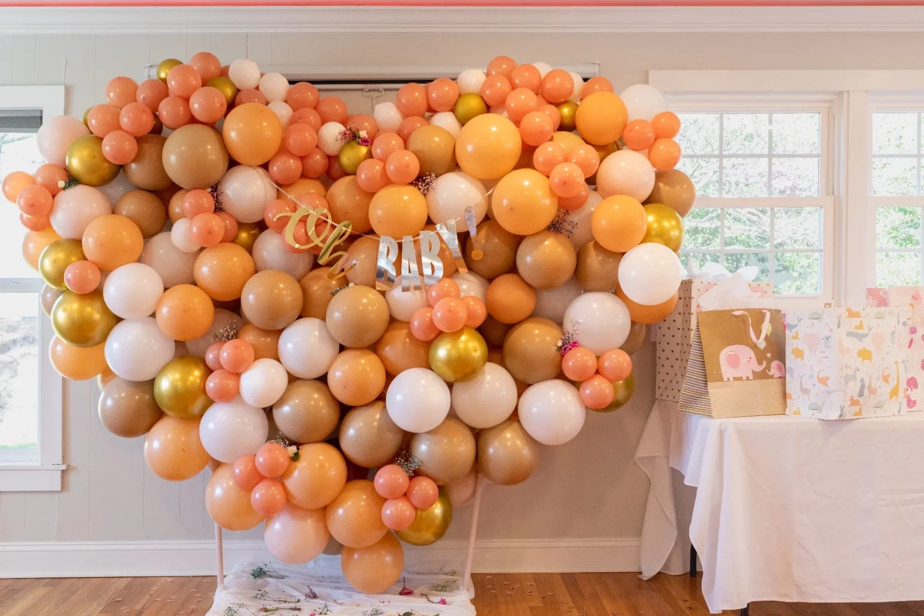 A heart-shaped balloon display with shades of orange, white, and gold, featuring a "BABY" banner. It is set against a wall with windows. Nearby is a table with gift bags, including one with an elephant design. Huntersville event venue