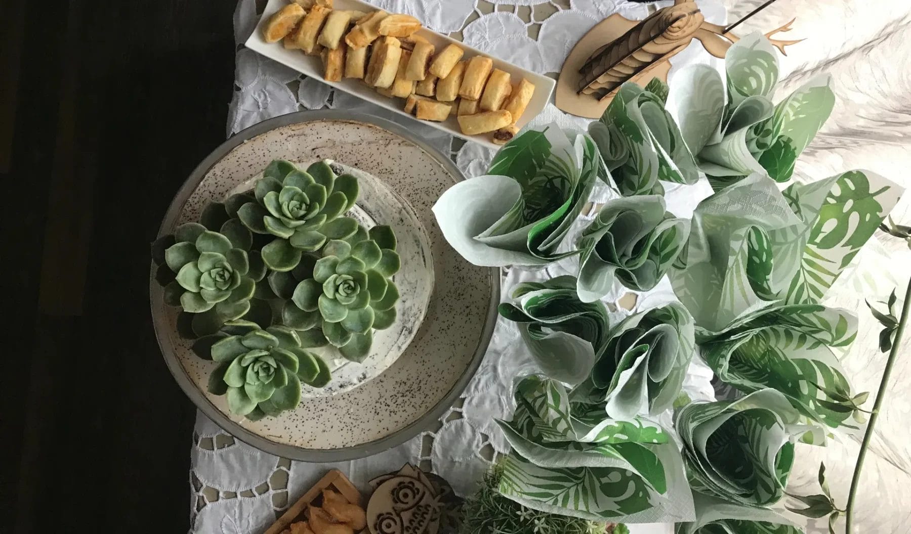 A top view of a decorative table setting featuring a centerpiece with succulents on a ceramic plate, snacks on elongated trays, and green leaf-patterned napkins folded neatly. All items are placed on a lace tablecloth. Huntersville event venue