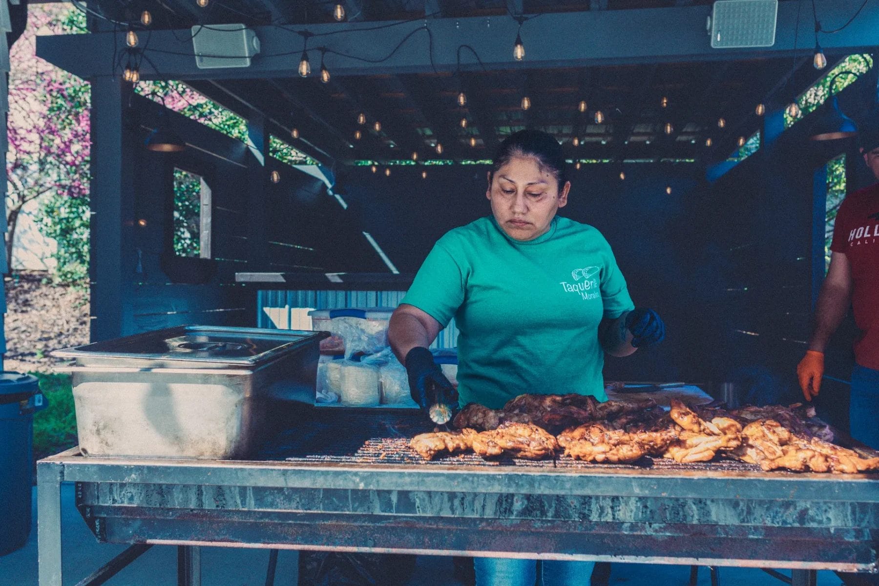 Woman in a teal shirt grilling meat on a large outdoor barbecue. She is focused on cooking under a canopy with string lights. Boxes and containers are around her, and there is greenery in the background. Huntersville event venue