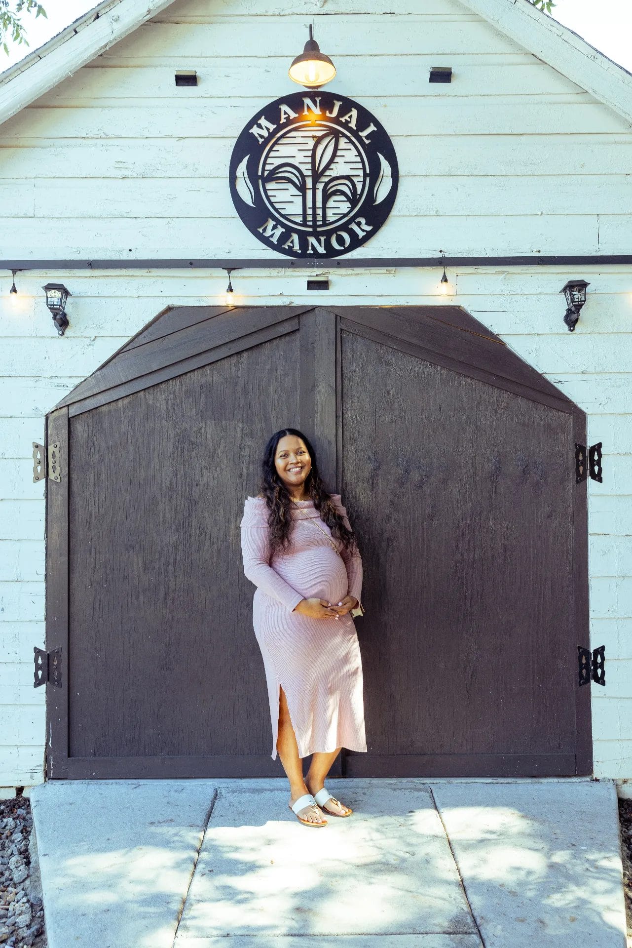 A pregnant woman in a lilac dress stands smiling in front of a large wooden door. Above the door is a sign that reads "Manjal Manor." The setting appears bright and outdoors. Huntersville event venue