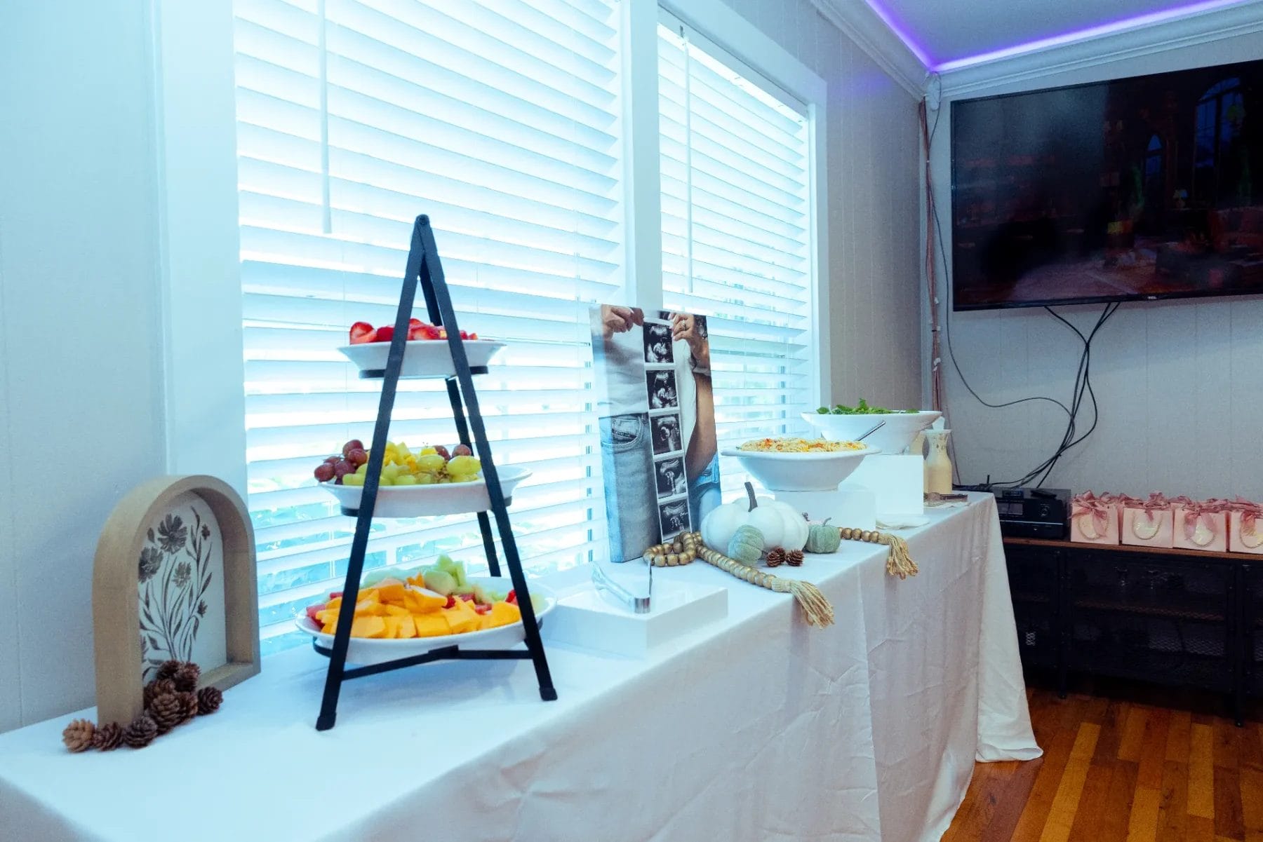 A brightly lit room with a buffet table. The table has a three-tiered stand with assorted fruits, a pumpkin, a bowl of popcorn, and decorative elements. A TV hangs on the wall, and pink bags are lined up below. Huntersville event venue