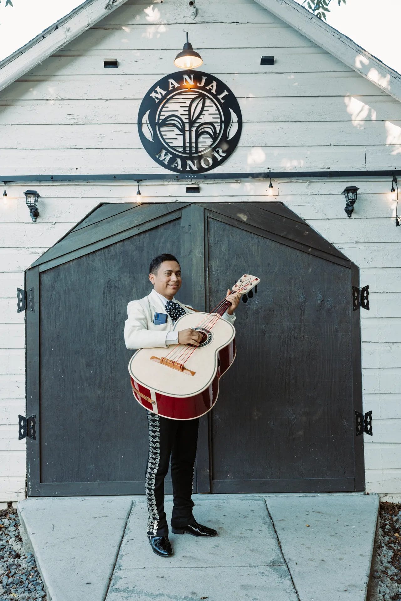 A Heartfelt Celebration at Manjal Manor: Great-Grandma’s 94th Birthday! 7 A person stands in front of a rustic building with dark wood doors, holding an oversized guitar. They are wearing a white jacket with intricate embroidery and black pants, under the "Manjal Manor" sign. The setting suggests a musical or festive occasion. Huntersville event venue