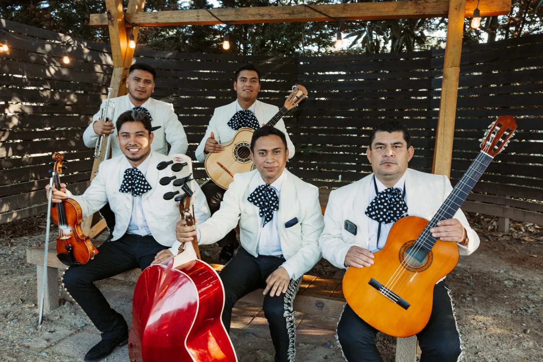 A mariachi band of five men in white jackets and black embroidered pants. They are holding instruments: a guitar, trumpet, violin, and guitarrón, sitting outdoors under a wooden structure with string lights. Wooden fence and trees in the background. Huntersville event venue