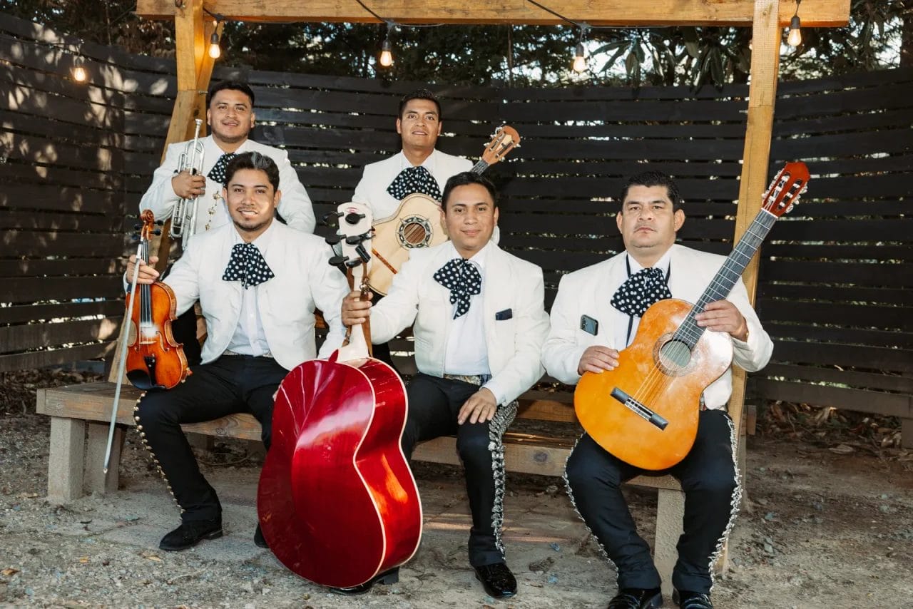 Five mariachi musicians in traditional attire pose outdoors, holding instruments including a violin, trumpet, guitarrón, vihuela, and guitar, under string lights on a wooden bench. Huntersville event venue