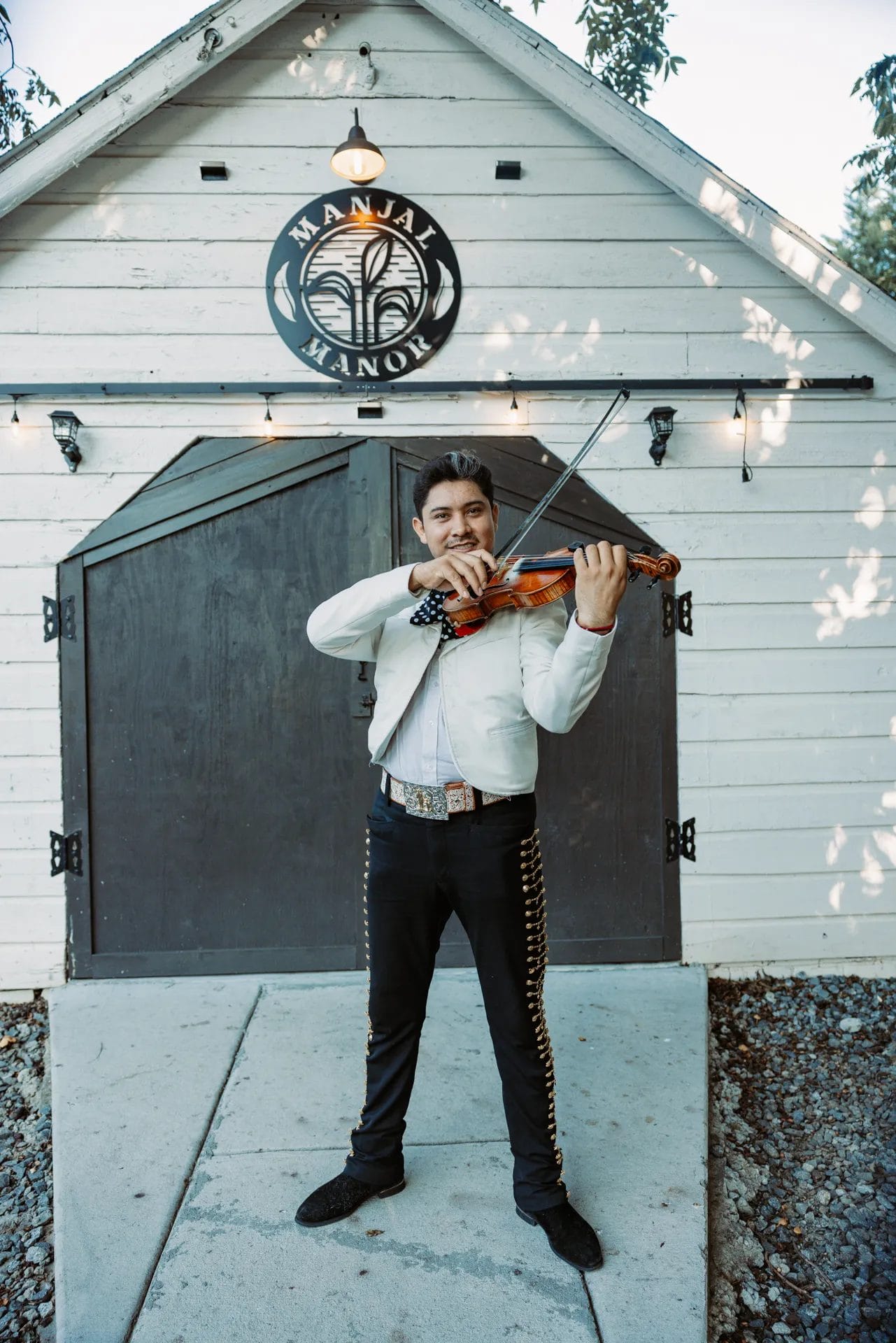 A Heartfelt Celebration at Manjal Manor: Great-Grandma’s 94th Birthday! 10 A man plays the violin while standing in front of a rustic white barn with dark wooden doors. He wears a white shirt and embroidered black pants. An emblem on the barn reads "Manual Manor." String lights decorate the building. Huntersville event venue