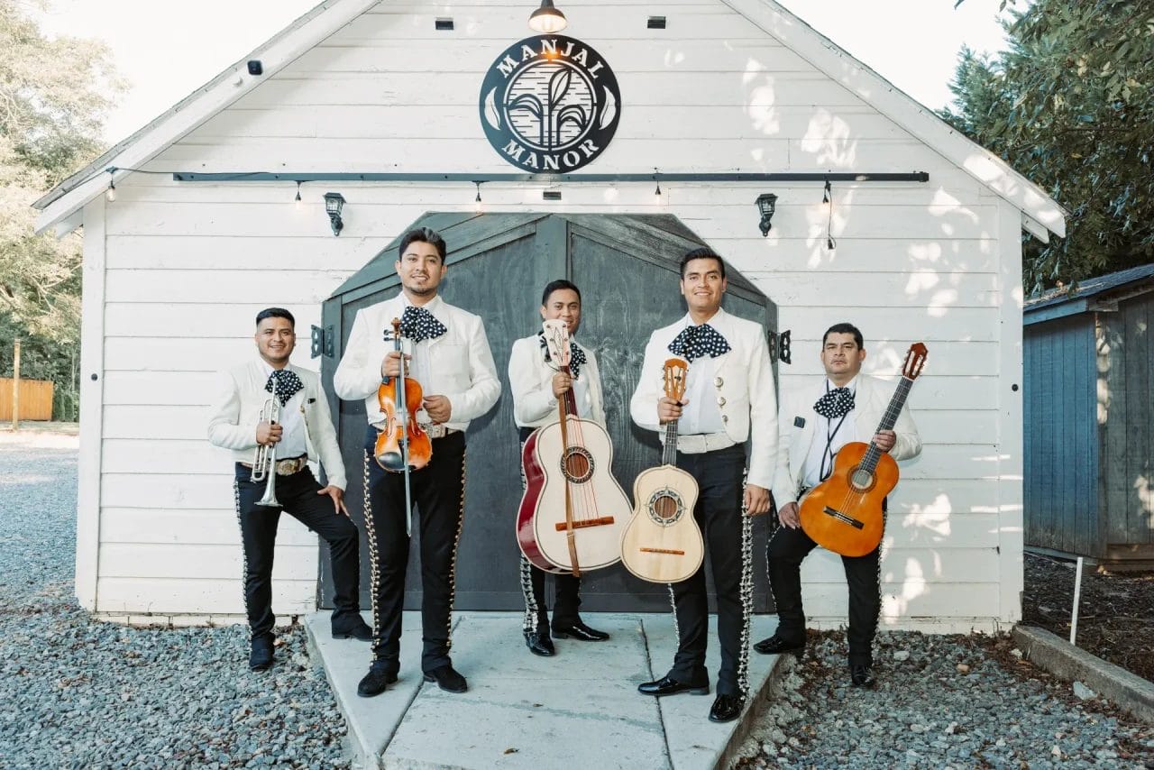 Five mariachi musicians stand in front of a white barn. They wear traditional charro outfits with embroidered jackets and hold various instruments, including violins, guitars, and a trumpet. The sign reads "Manja Manor. Huntersville event venue