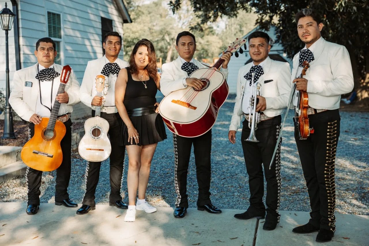 A woman stands smiling with a mariachi band. The group consists of five male musicians wearing traditional white uniforms with black pants and bows. Instruments include guitars, a guitarrón, a trumpet, and a violin. They're outdoors near a building. Huntersville event venue