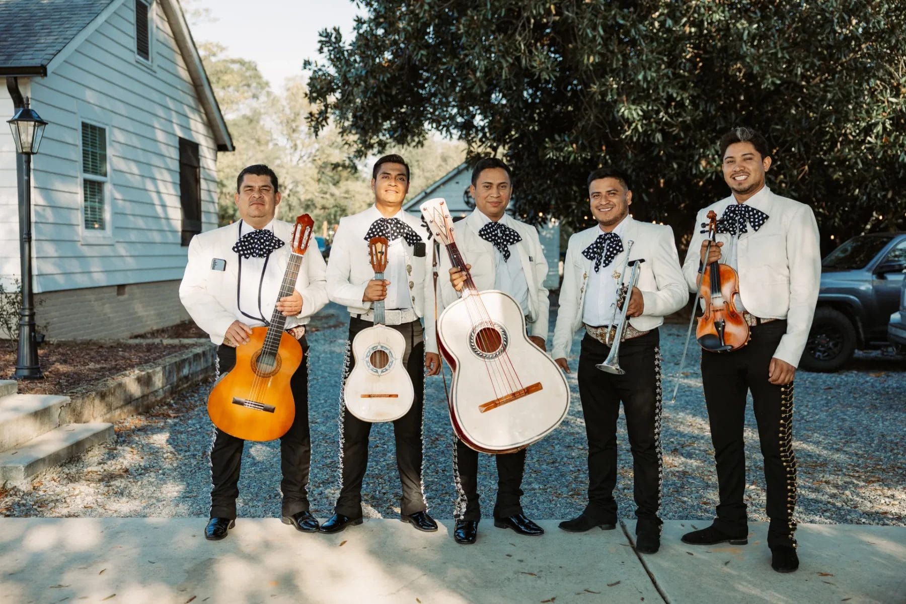 Five mariachi musicians stand outdoors holding instruments, including guitars, a trumpet, and a vihuela. They are dressed in traditional charro suits with intricate embroidery and black bow ties. A house and trees are visible in the background. Huntersville event venue