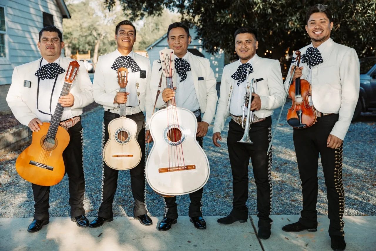 Five mariachi musicians stand smiling, wearing traditional outfits with bow ties. They hold various instruments, including guitars, a trumpet, a violin, and a vihuela, on a sunny day with trees in the background. Huntersville event venue