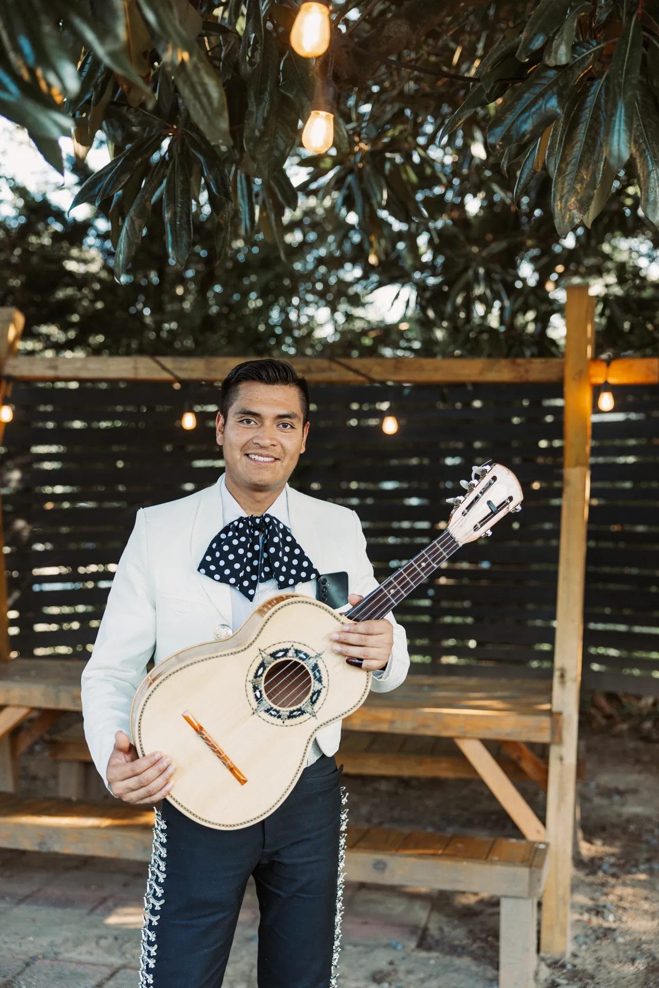 A Heartfelt Celebration at Manjal Manor: Great-Grandma’s 94th Birthday! 8 A man in a white jacket and black pants holds a guitar while standing outdoors. He is under string lights, with a wooden bench and greenery in the background. Huntersville event venue