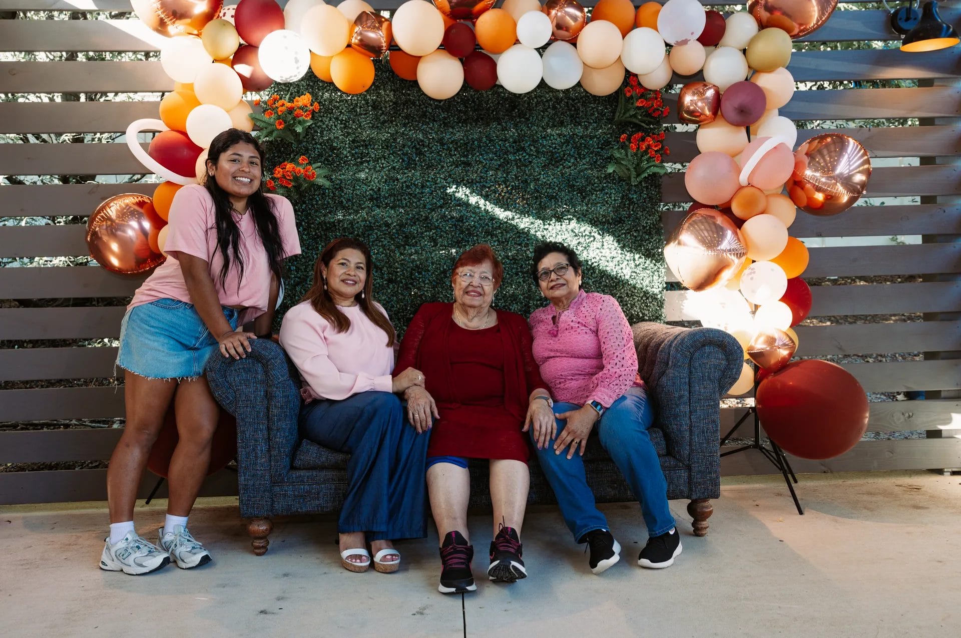 A group of four women posing together. Three are seated on a couch while one stands beside them. Behind them, a backdrop decorated with balloons in shades of orange, red, and white. The setting appears to be a celebratory event. Huntersville event venue