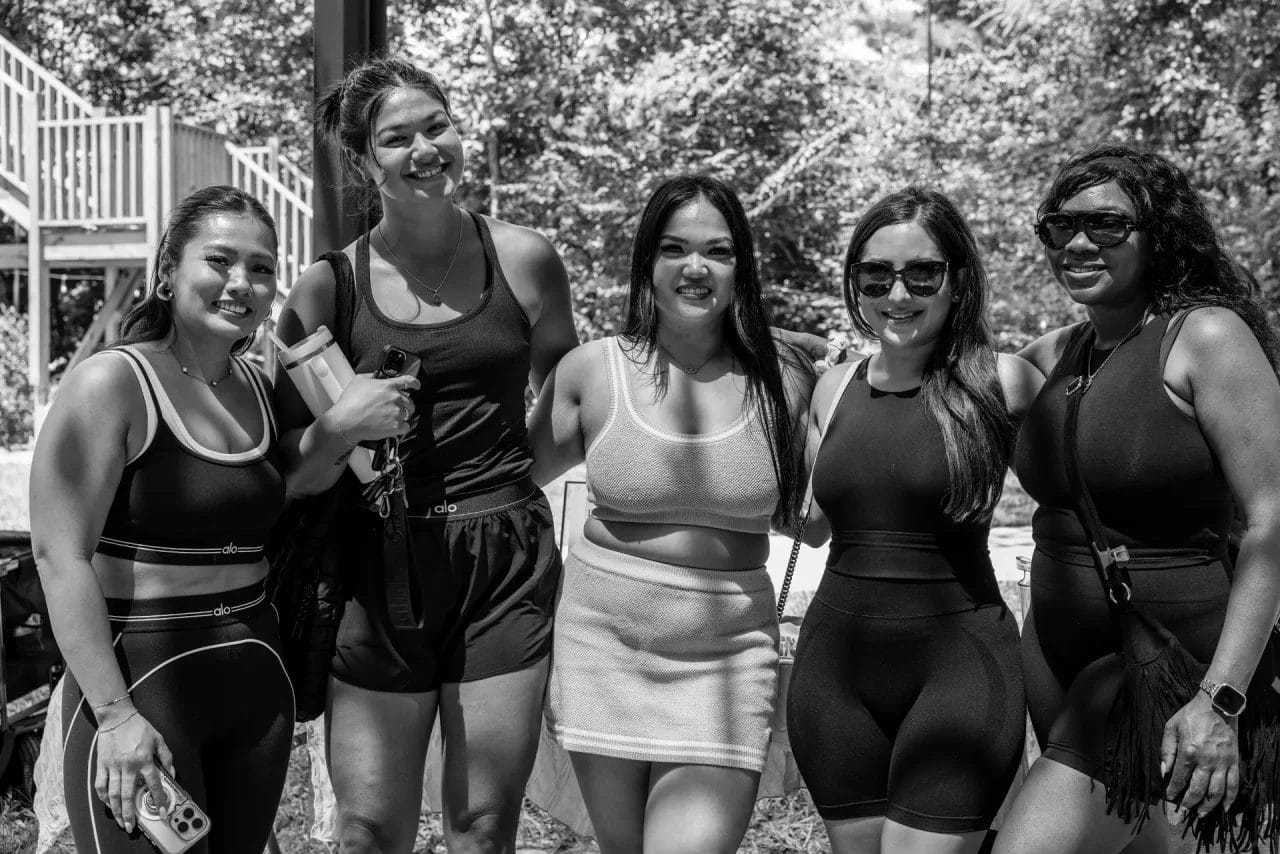 Five women stand close together outdoors, smiling at the camera. They are wearing athletic outfits. Trees and a wooden structure are in the background. The image is in black and white. Huntersville event venue