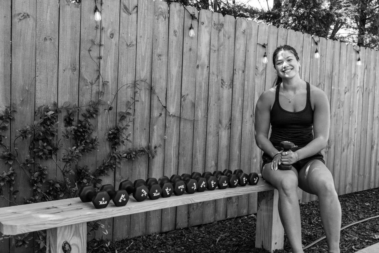 A woman in workout clothes sits on a bench outdoors, smiling and holding a dumbbell. A row of dumbbells is lined up on the bench beside her, with a wooden fence and string lights in the background. Huntersville event venue