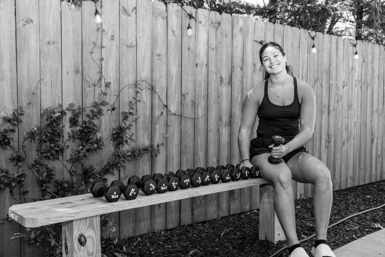 A woman in athletic wear sits on a bench outdoors, smiling, with a row of dumbbells beside her. She holds one weight in her hand. The setting is a fenced backyard with string lights and some greenery. Black and white image. Huntersville event venue