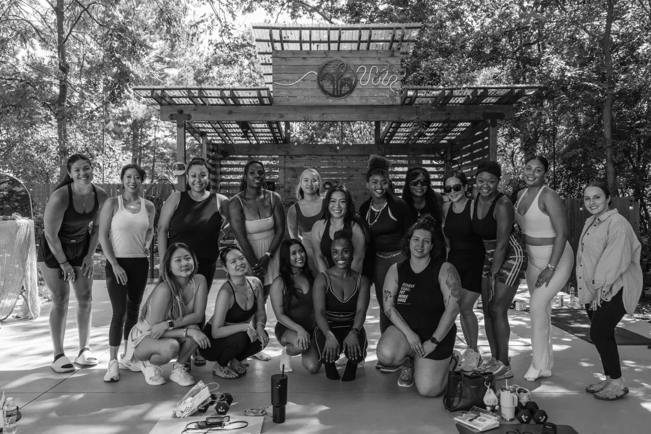 A group of women in activewear pose together outdoors in front of a wooden shelter, smiling at the camera. Sunlight filters through trees and the structure behind them. Fitness gear is visible on the ground. Huntersville event venue