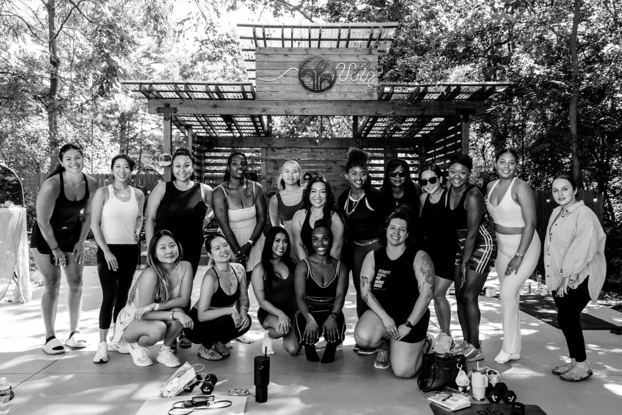 A group of people in athletic wear pose together outdoors in front of a wooden structure, smiling at the camera. Some are standing, others are kneeling, and exercise equipment is on the ground. Huntersville event venue