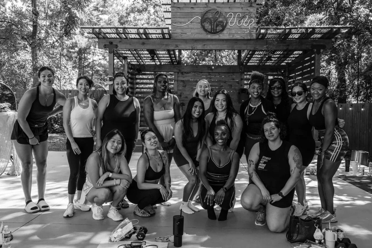 A group of sixteen women pose and smile outdoors on a concrete patio, under a wooden pergola, surrounded by trees. Most wear athletic clothing. The photo is black and white. Huntersville event venue