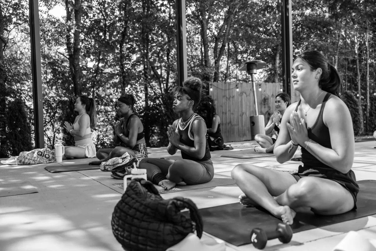 A group of women sit cross-legged on yoga mats outdoors, meditating with hands in prayer position. Sunlight filters through trees in the background, creating a calm and peaceful atmosphere. Huntersville event venue