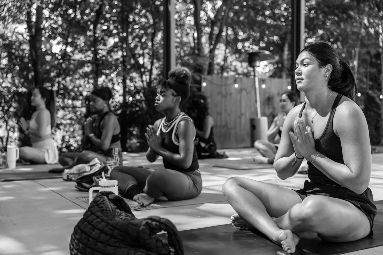 A group of people sit cross-legged on yoga mats outdoors, eyes closed and hands in prayer position, meditating together under natural light with trees and greenery in the background. Huntersville event venue