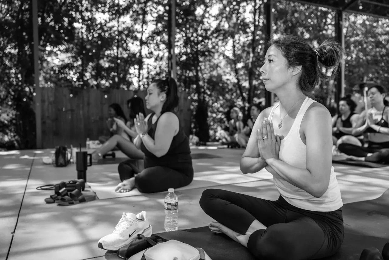 A group of women sit cross-legged on yoga mats outdoors, hands in prayer position, participating in a yoga session. Trees and sunlight are visible in the background. Yoga accessories and water bottles are nearby. Huntersville event venue
