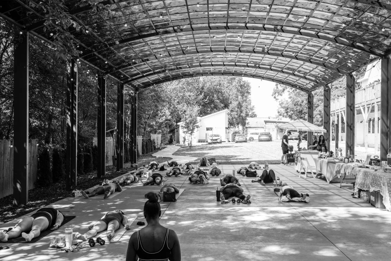 A black and white photo of a group yoga class under a large, open-air pavilion. People lie on mats in a relaxed pose, while an instructor stands at the front. Tables and parked cars are visible in the background. Huntersville event venue