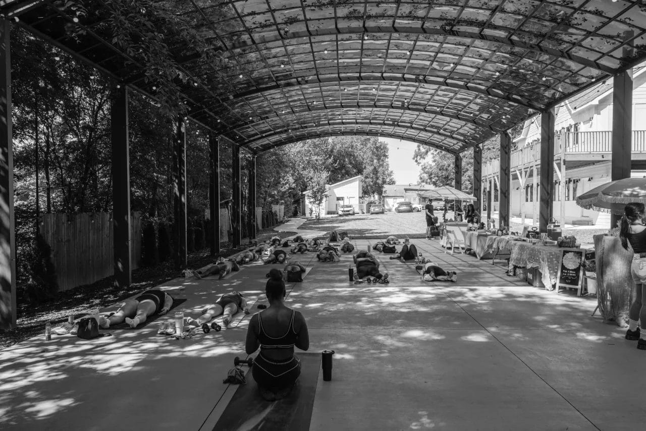 A group of people practice yoga outdoors under a large arched canopy, while market stalls with tables are set up to the right; sunlight filters through the overhead structure. Huntersville event venue