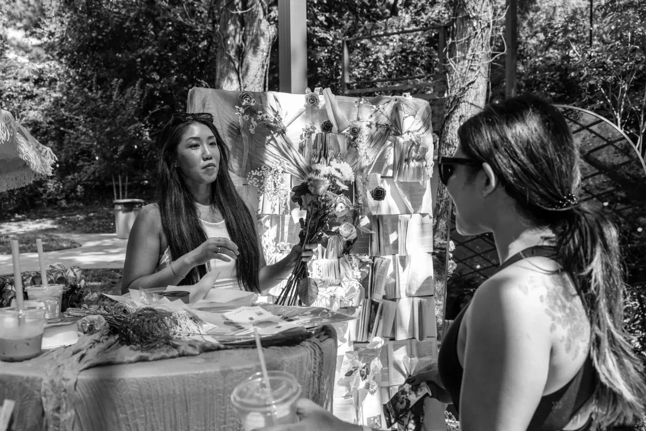 A woman arranges a bouquet of flowers at an outdoor market booth, while another woman with sunglasses stands across from her holding a drink. Behind them are hanging floral decorations. The scene is in black and white. Huntersville event venue