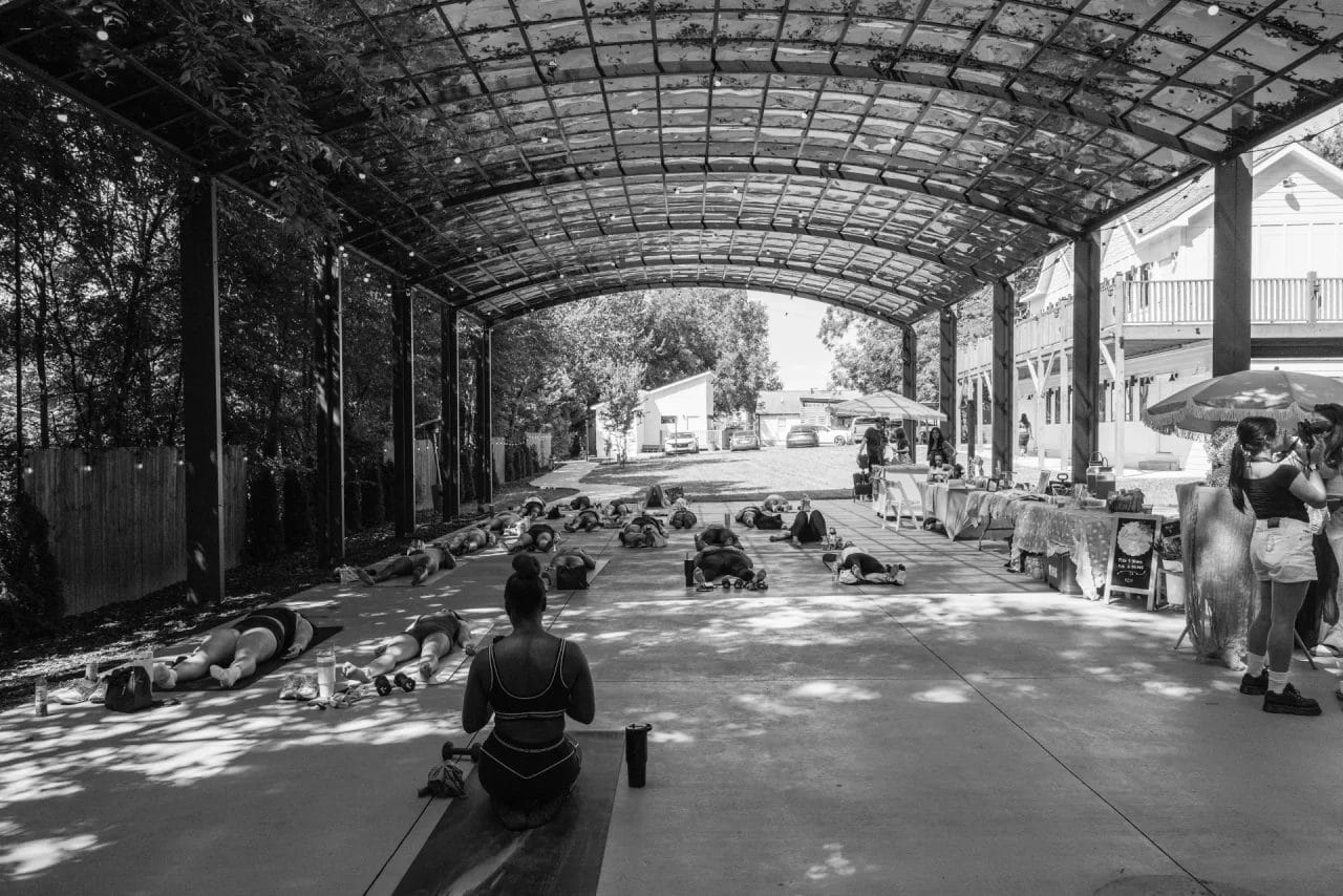 A group of people practice yoga under a large covered outdoor pavilion, with market stalls and onlookers nearby. Sunlight filters through the roof, casting patterned shadows on the ground. Huntersville event venue