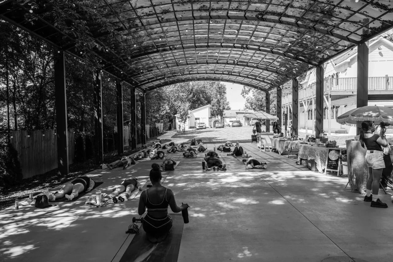 A group of people participates in a fitness or yoga class under a large, open-sided metal pavilion; tables with items are set up on the right, and sunlight filters through the roof. Huntersville event venue