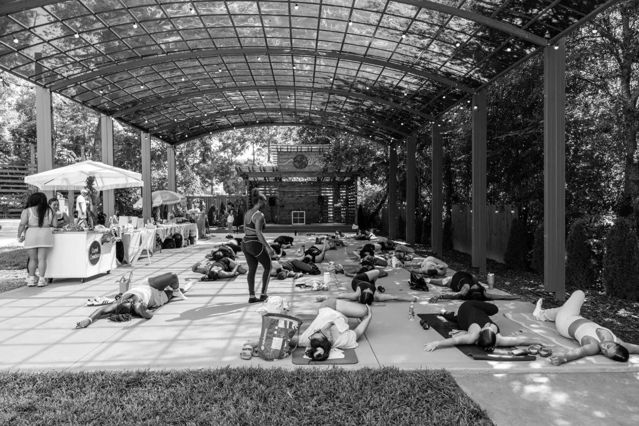 A group of people practice yoga on mats under a large, open-air pavilion. An instructor stands among them, while a few people are at tables on the side. The scene is calm and shaded, surrounded by trees. Huntersville event venue