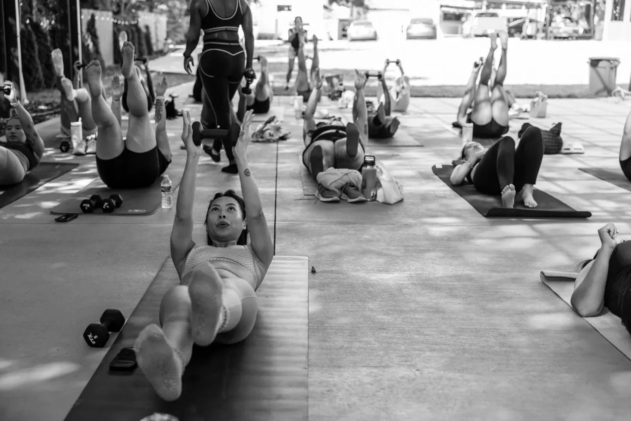 A group of people exercise on yoga mats outdoors, lying on their backs with legs raised and arms extended upward, following an instructor at the front. The scene is in black and white. Huntersville event venue