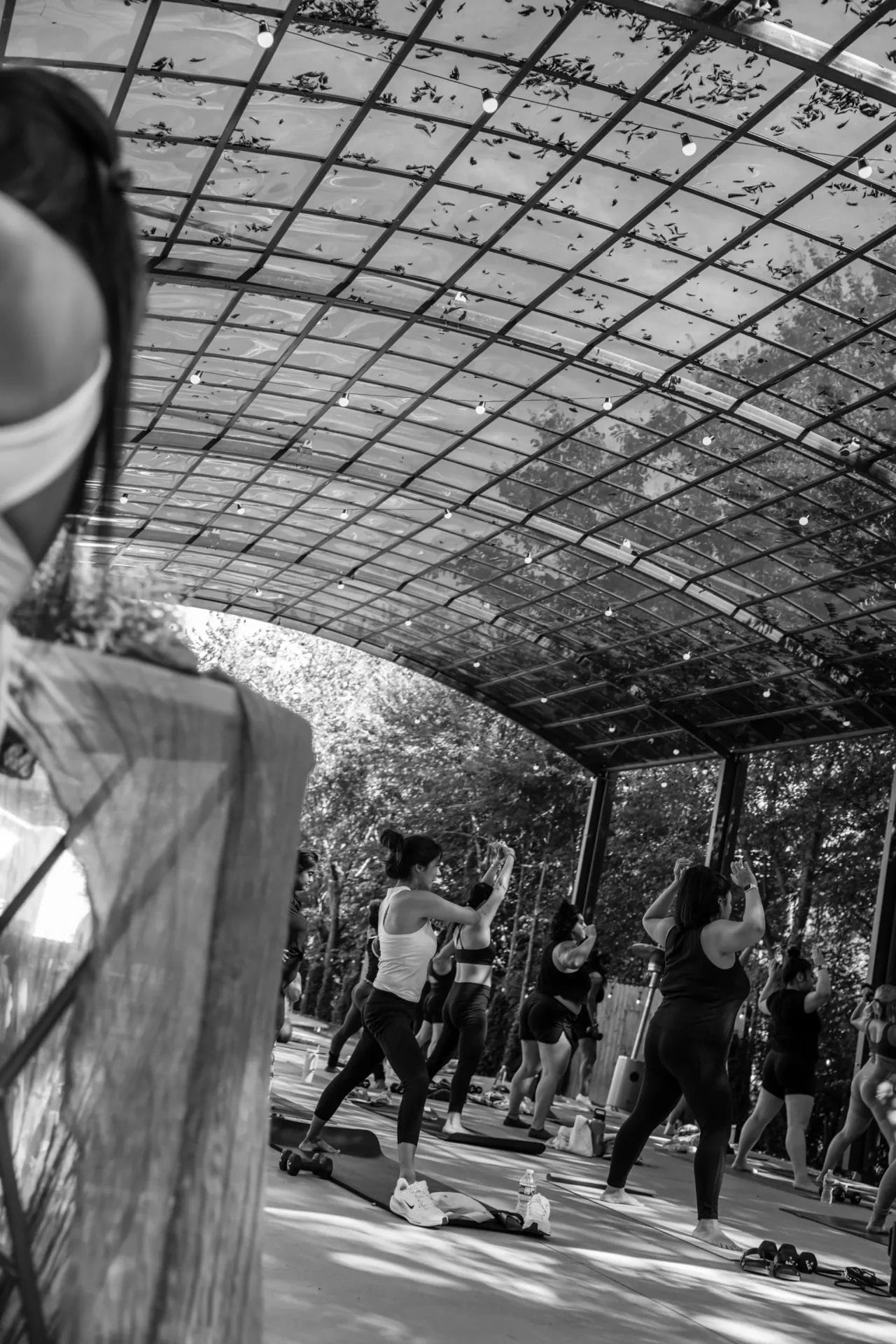 A group of people participate in an outdoor fitness class under a large, open shelter with a glass roof, surrounded by trees. Some are stretching, while others follow along with the instructor. Huntersville event venue