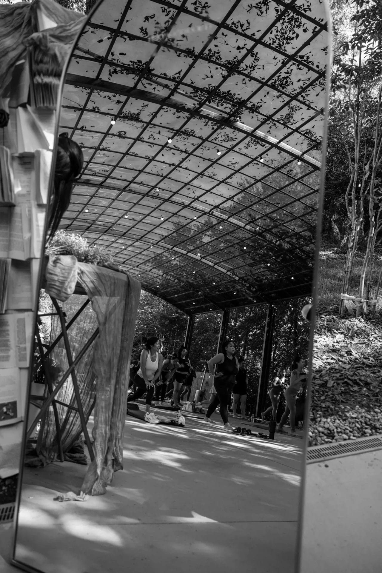 A group of people exercise in an open-air pavilion surrounded by trees, reflected in a large mirror decorated with hanging fabric and books. The scene is in black and white. Huntersville event venue