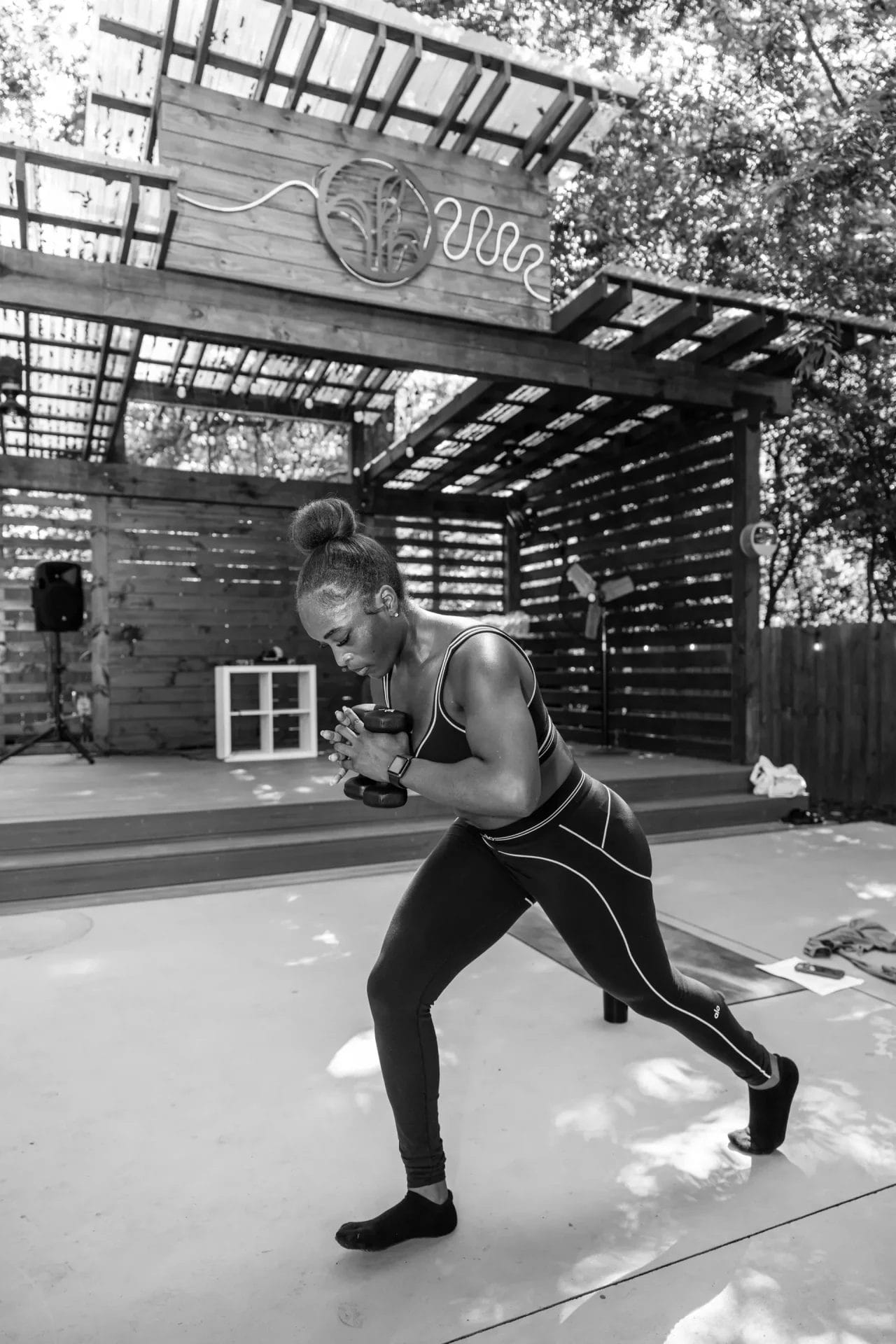 A woman in athletic wear exercises outdoors on a concrete surface, holding a dumbbell with both hands. She is in a lunge position. A wooden stage structure with a decorative symbol stands behind her. The image is in black and white. Huntersville event venue