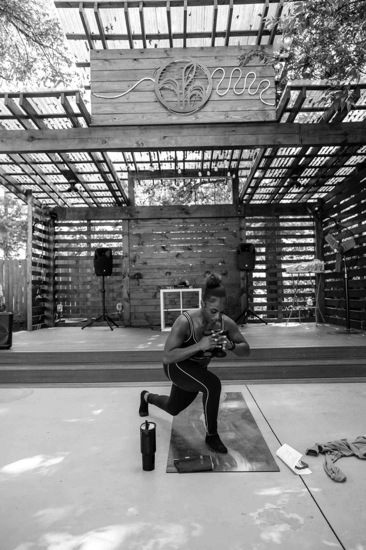 A woman in athletic wear performs a lunge exercise on a mat outdoors under a wooden pavilion. Exercise equipment, a water bottle, and a towel are nearby. The scene is in black and white. Huntersville event venue