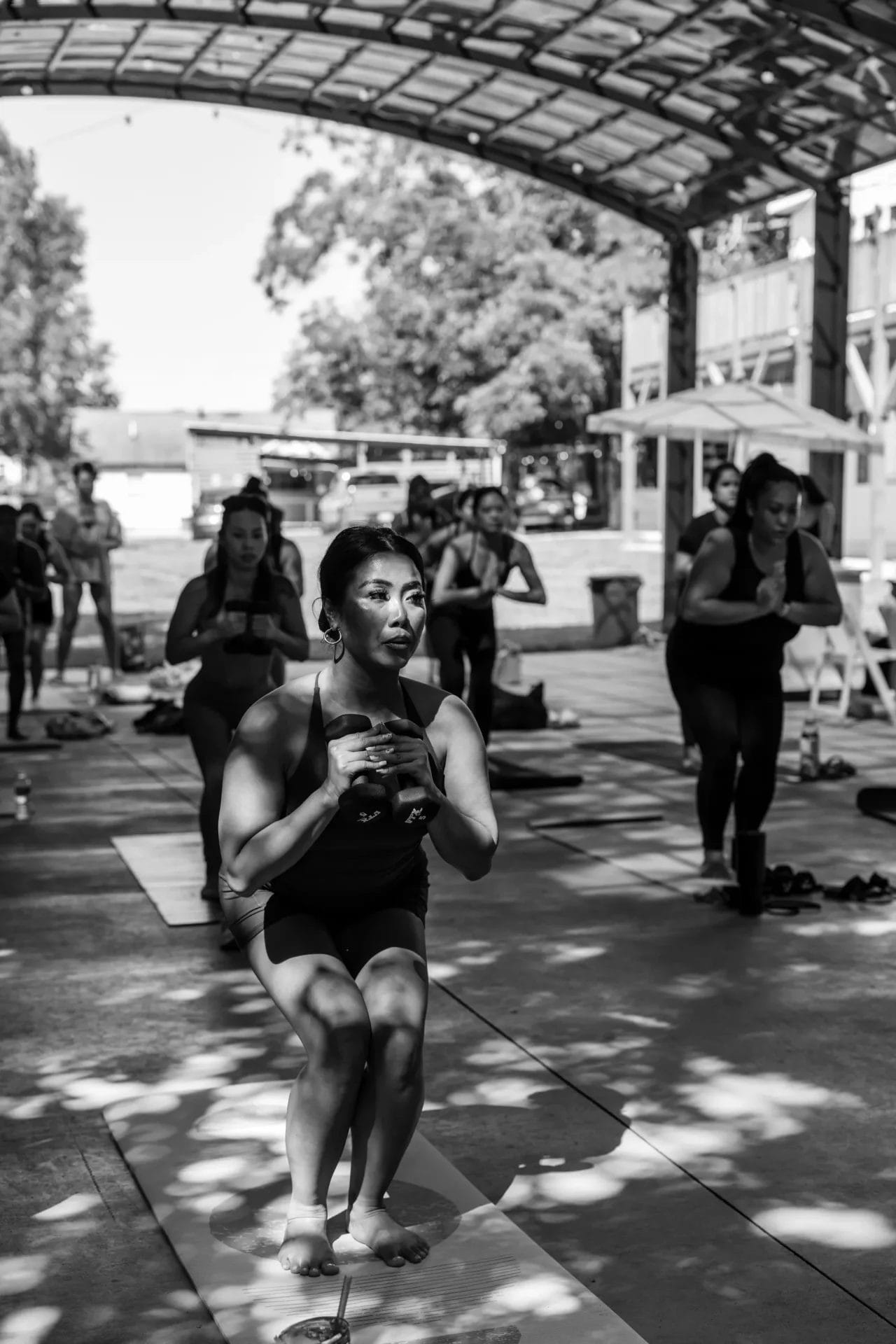 A group of people practice yoga together outdoors under a large, open-roof pavilion. The main focus is a woman in front, balancing in a squat position on her yoga mat. The scene is in black and white. Huntersville event venue