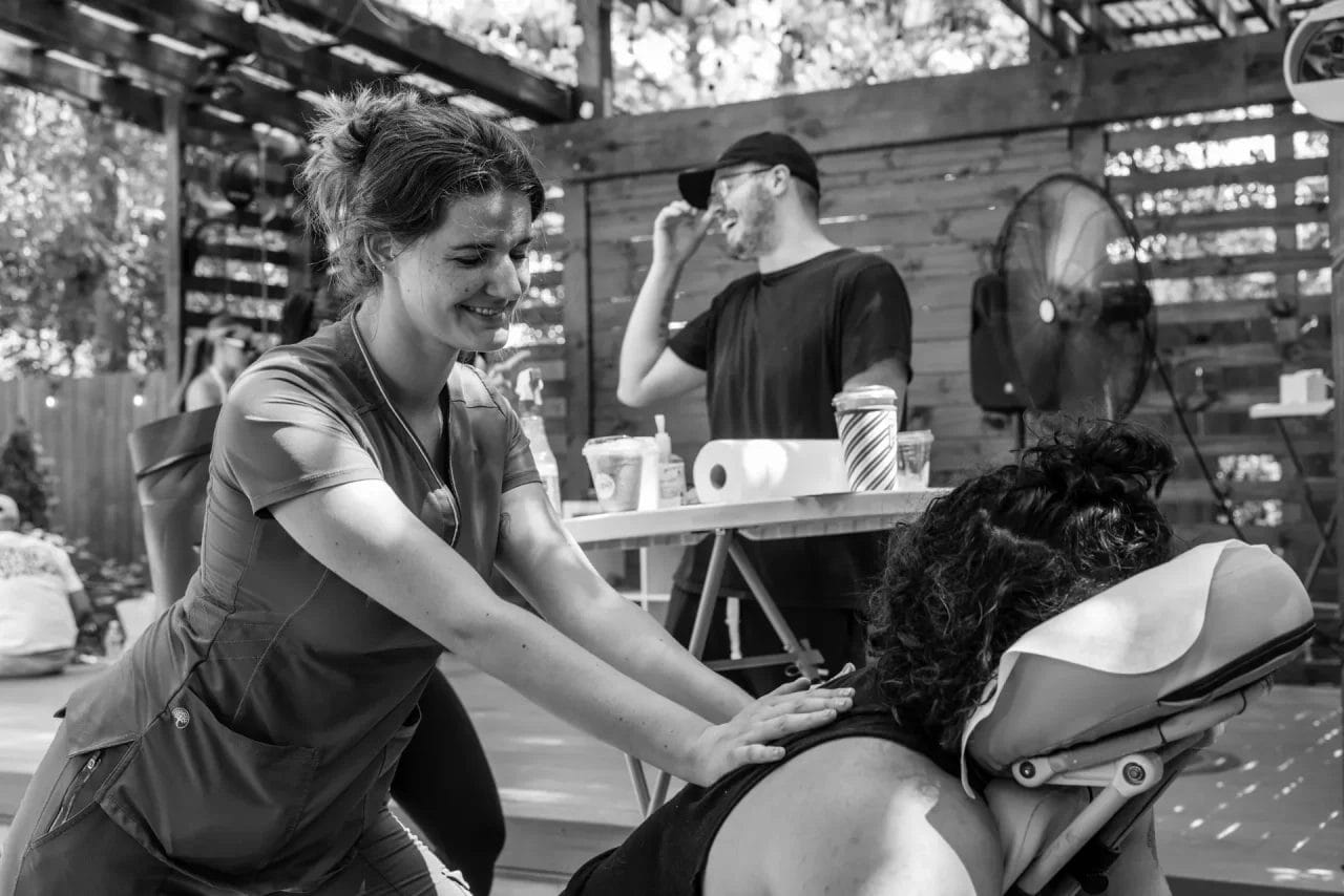 A woman gives a seated back massage to another woman outdoors, both smiling. In the background, a man in a cap stands by a table with drinks, laughing. The setting appears relaxed and social. Huntersville event venue