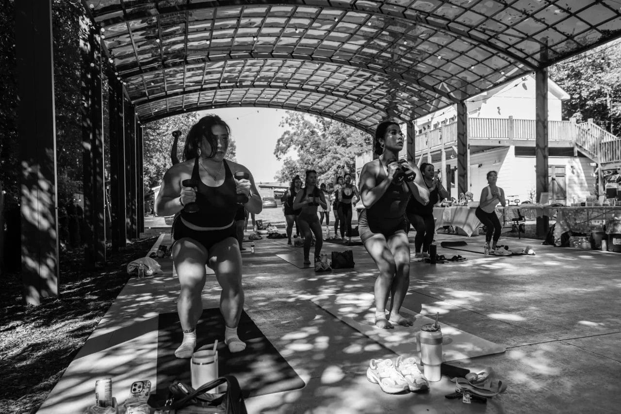 A group of people participate in an outdoor exercise class under a large open-air pavilion, performing squats on yoga mats in athletic wear. Sunlight filters through the structure, casting dappled shadows. Huntersville event venue