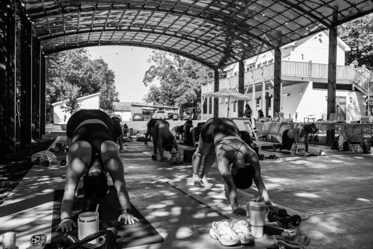People practicing yoga outdoors under a large open-air structure, performing downward dog poses on yoga mats. Water bottles and small weights are nearby; sunlight filters through the roof above. The image is in black and white. Huntersville event venue